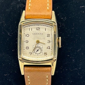 May include: A gold-toned wristwatch with a white face and black hands. The watch has a brown leather band with white stitching. The face of the watch has black Roman numeral hour markers and the word "HARMAN" printed in black.