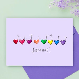 Puede incluir: Una tarjeta de felicitación de color violeta claro con una fila de notas musicales en forma de corazón en rojo, rosa, morado, naranja, verde, amarillo, azul y violeta. Debajo, el texto escrito a mano dice: "Just a note!"