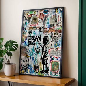 May include: Framed art print featuring a graffiti-style design with the words "DREAM BIG" in blue and black. A silhouette of a child painting a heart is visible. The artwork includes various symbols and text elements, with a black frame.