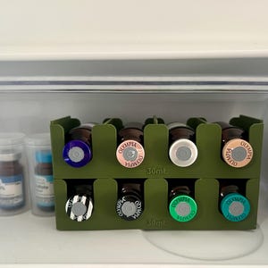 May include: A green organizer holds eight small brown glass bottles with colorful caps. The caps are silver, pink, blue, green, and black and white striped. The bottles are labeled "Olympia" and "30ml."