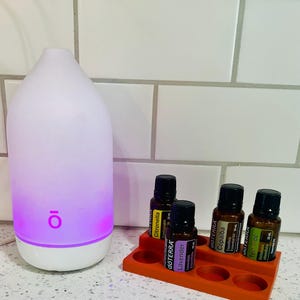 May include: A white teardrop-shaped essential oil diffuser with a glowing purple base and the logo "ō" on the front, accompanied by an orange plastic stand holding four 15 ml dōTERRA essential oil bottles labeled Citronella, Lavender, Copaiba, and Tea Tree.