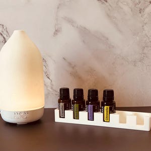 May include: A white, teardrop-shaped essential oil diffuser with a glowing light base, positioned next to a white rectangular organizer tray holding four amber glass essential oil bottles labeled Copaiba, Tea Tree, Lavender, and Citronella.