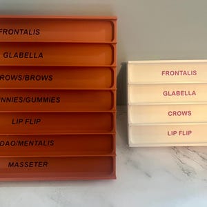 Könnte beinhalten: Zwei Sätze rechteckiger, gestufter Tabletts. Ein Satz ist orange mit schwarzen Textetiketten: Frontalis, Glabella, Crows/Brows, Bunnies/Gummies, Lip Flip, Dao/Mentalis und Masseter. Der andere Satz ist weiß mit rosa Textetiketten: Frontalis, Glabella, Crows und Lip Flip.