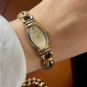 Puede incluir: Un reloj de pulsera ovalado de dos tonos, dorado y plateado, con una esfera blanca y marcadores de hora en números romanos negros. El reloj tiene una correa dorada con un diseño de eslabones único.