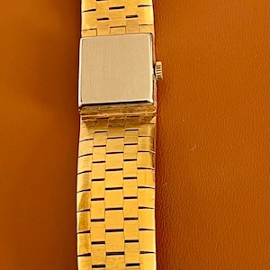 May include: Gold-toned watch band with a rectangular watch face. The band is made of small, rectangular links. The watch face is silver-toned. The band is displayed on a brown surface.