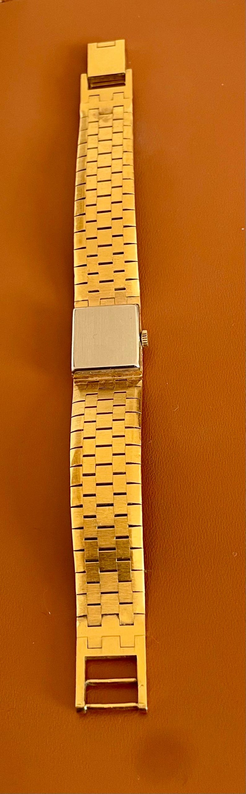 May include: Gold-toned watch band with a rectangular watch face. The band is made of small, rectangular links. The watch face is silver-toned. The band is displayed on a brown surface.