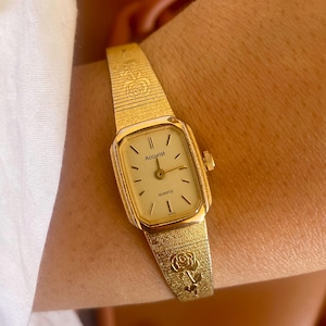 Op de afbeelding: Een goudkleurig Accurist Quartz horloge met een rechthoekige wijzerplaat en een gestructureerde band. De wijzerplaat is crèmekleurig met zwarte markeringen en wijzers. De band heeft een rozenontwerp. Een klassiek accessoire.