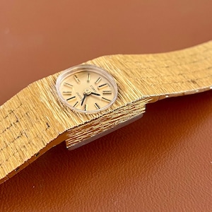 May include: A gold-toned wristwatch with a textured band and a round face. The watch has a light-colored dial with black hands and markers. The band appears to be made of a woven metal material.