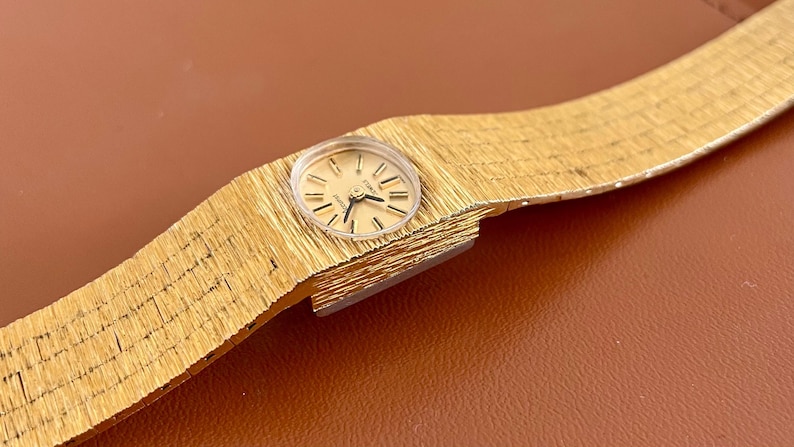 May include: A gold-toned wristwatch with a textured band and a round face. The watch has a light-colored dial with black hands and markers. The band appears to be made of a woven metal material.