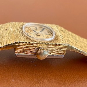 May include: A vintage gold-toned wristwatch with a textured band and a round face. The watch face features Roman numeral hour markers and delicate hands. The band appears to be made of woven metal. The watch is displayed on a brown surface.