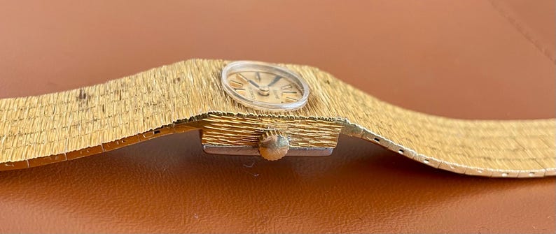 May include: A vintage gold-toned wristwatch with a textured band and a round face. The watch face features Roman numeral hour markers and delicate hands. The band appears to be made of woven metal. The watch is displayed on a brown surface.