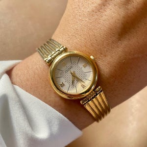 May include: A gold-toned wristwatch with a white face and a diamond-like pattern. The watch has Roman numeral hour markers and the word "Leore" printed on the face.
