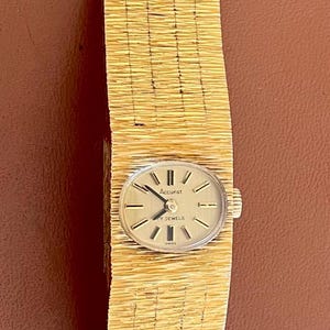 May include: A vintage gold-toned wristwatch with a textured band. The watch face is oval-shaped with black hands and markers. The band has a woven appearance. The watch is set against a brown background.
