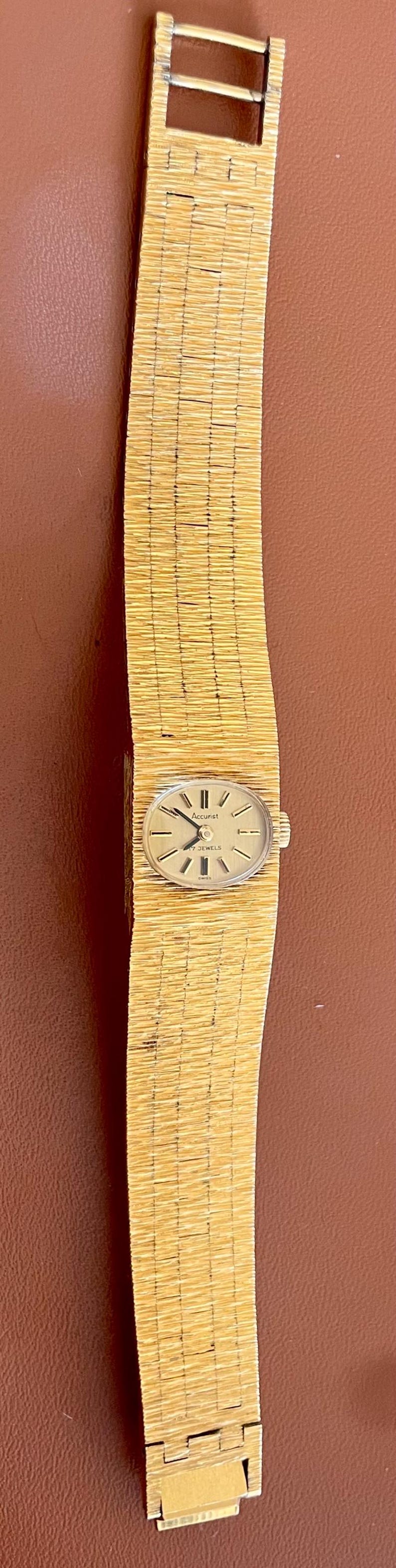 May include: A vintage gold-toned wristwatch with a textured band. The watch face is oval-shaped with black hands and markers. The band has a woven appearance. The watch is set against a brown background.