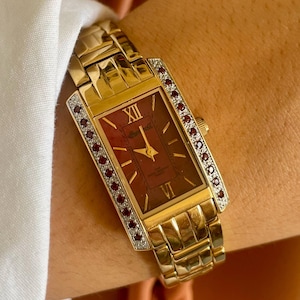 May include: A gold-tone rectangular wristwatch featuring a deep red dial with gold hands and Roman numeral markers at twelve and six o'clock. The bezel is accented with two vertical rows of small, sparkling clear stones and red gems.