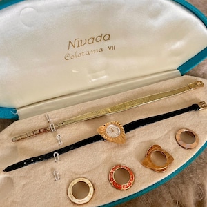 May include: Vintage Nivada Colorama VII watch set in a teal and cream case. The set includes a gold-toned watch with a black band, a gold-toned band, and several interchangeable gold-toned bezels with red and cream accents.