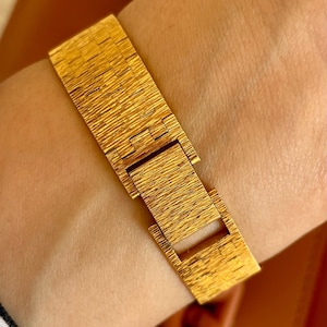 May include: A gold-colored bracelet with a textured, linear pattern. The bracelet is fastened with a rectangular clasp. The jewelry is worn on a wrist. The bracelet appears to be made of metal.