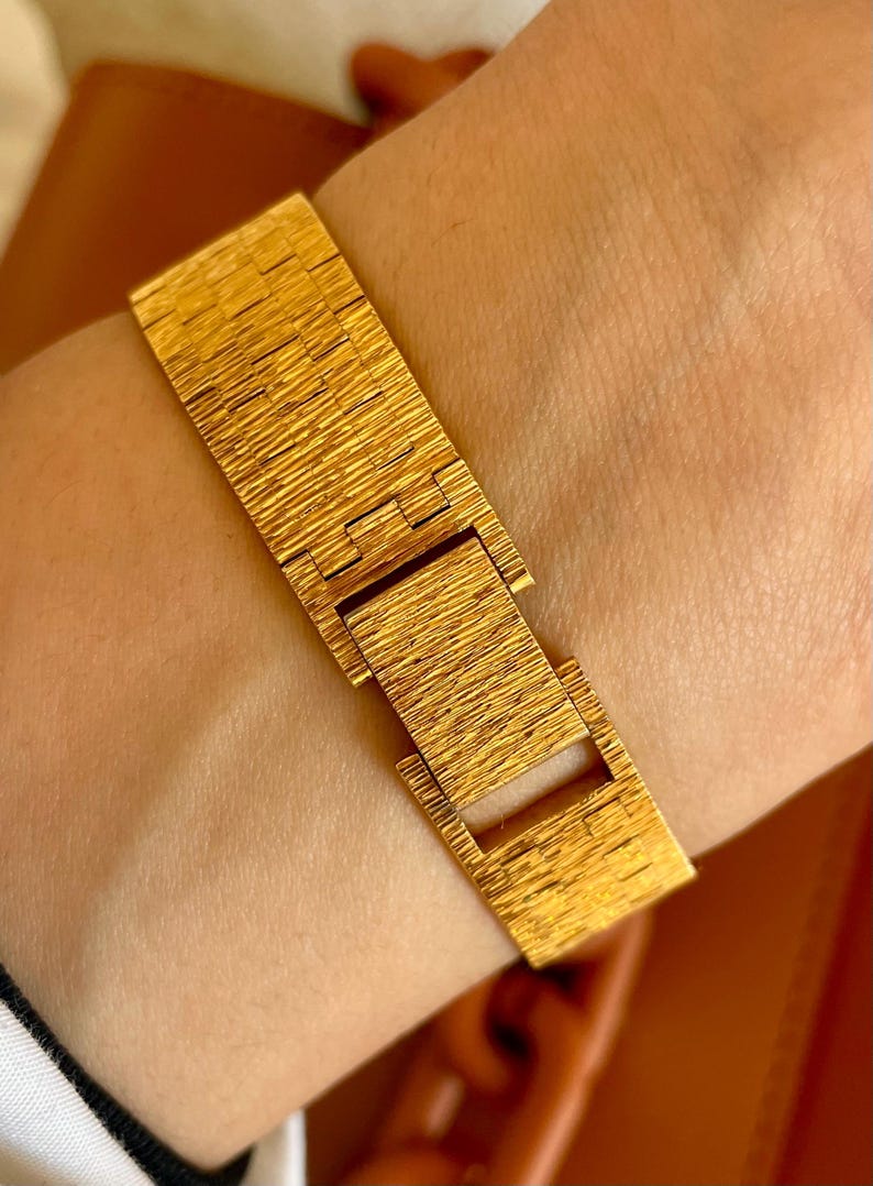 May include: A gold-colored bracelet with a textured, linear pattern. The bracelet is fastened with a rectangular clasp. The jewelry is worn on a wrist. The bracelet appears to be made of metal.
