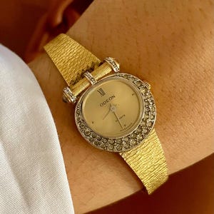 May include: A gold-toned wristwatch with a textured band and a round face. The watch face is surrounded by small, clear stones. The watch face displays the word "ODEON" and "QUARTZ". The band is approximately 0.5 inches wide.