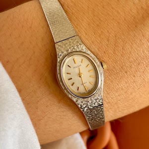 May include: A silver-tone Pulsar quartz watch with an oval face, a light-coloured dial, and gold-tone hands and hour markers. The watch features a textured metal link band with a cross-hatch pattern and a small gold-tone crown on the side.