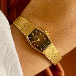 Pode incluir: Um relógio Rotary dourado com um mostrador retangular escuro e ponteiros dourados. O relógio tem uma pulseira dourada texturizada e é usado no pulso. O mostrador mostra o nome da marca.