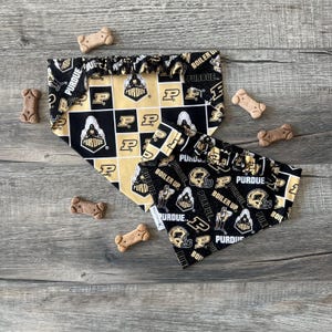 May include: Two Purdue University dog bandanas in black and gold, with the Purdue logo and text. The bandanas are surrounded by dog bone-shaped treats on a wooden surface. The bandanas have a checkered and patterned design.