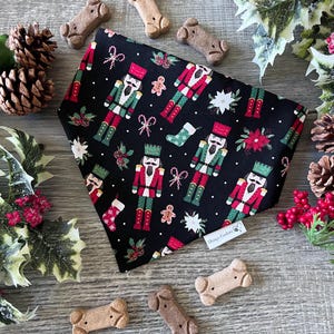 May include: A black triangular pet bandana featuring a repeating pattern of nutcrackers, gingerbread men, candy canes, stockings, and poinsettias. A small white fabric tag with the text "Chrissy's Creations" is attached to the lower edge.