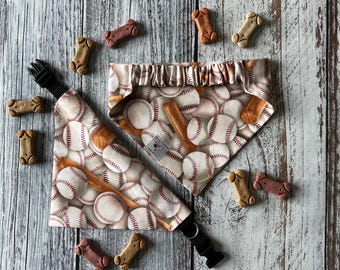May include: A baseball-themed dog bandana and collar set with a pattern of baseballs and bats. The set includes a bandana and a collar with a black buckle. Several dog biscuits are scattered around the set on a wooden surface.
