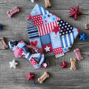 May include: A patriotic dog bandana and bow tie set with a patchwork design in red, white, and blue. The bandana and bow tie feature stars and stripes patterns. Decorative stars and dog bone treats surround the set on a wooden surface.
