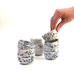 Könnte beinhalten: Eine Reihe kleiner, weißer Keramikschalen mit blauem Sprenkelmuster. Die Schalen sind gestapelt, eine wird von einer Hand gehalten. Die Schalen sind rund und eignen sich zum Servieren kleiner Portionen. Jede Schale hat einen Durchmesser von ca. 10 cm.