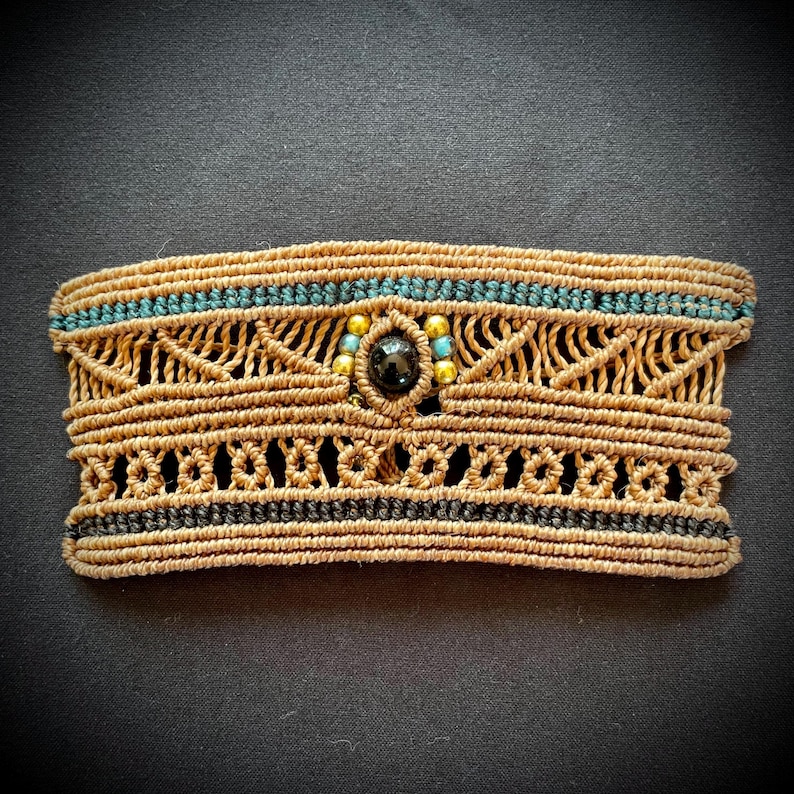 May include: A brown macrame bracelet with a black and turquoise design. The bracelet has a black gemstone in the center surrounded by gold beads.