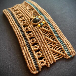 May include: A brown woven bracelet with black and blue accents. The bracelet has a black gemstone and gold beads in the center.