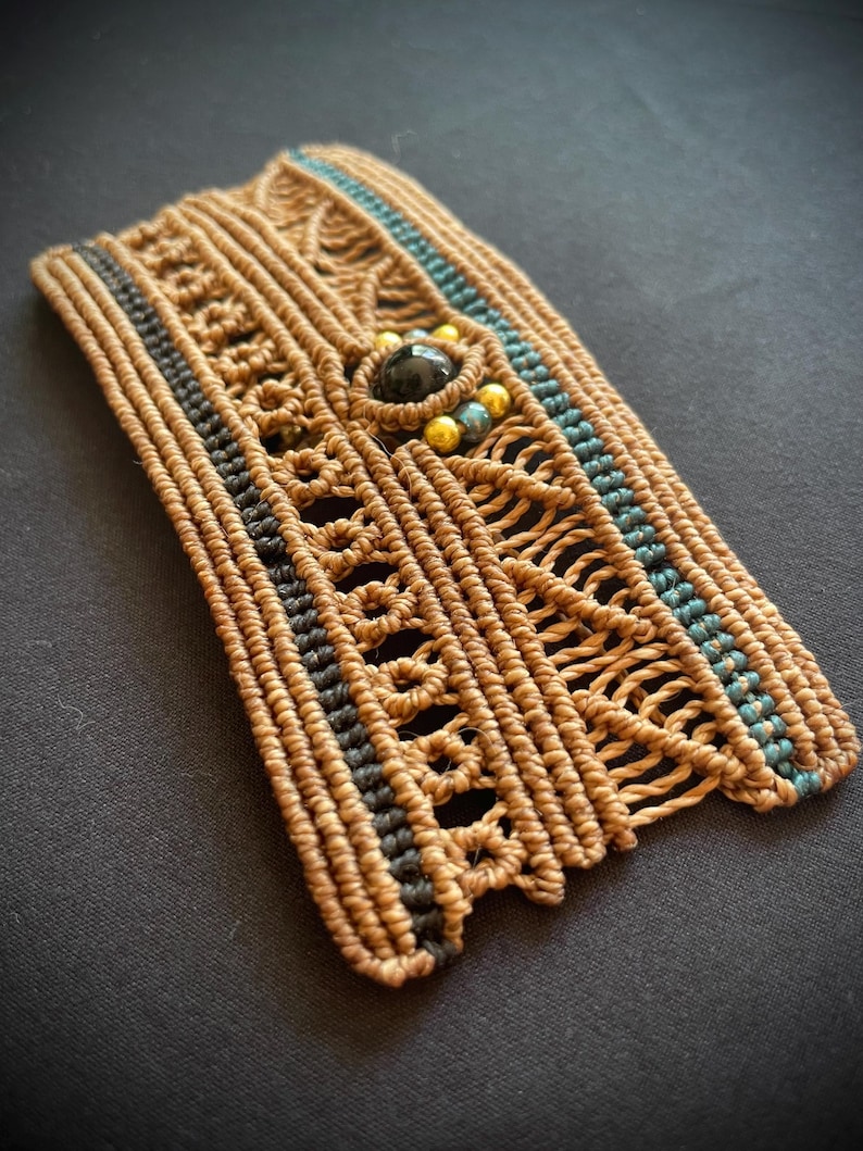 May include: A brown woven bracelet with black and blue accents. The bracelet has a black gemstone and gold beads in the center.