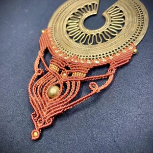 May include: A handcrafted necklace featuring a large, ornate, bronze-colored pendant with intricate cut-out designs. The pendant is connected to a macrame-style, reddish-brown woven section with a central bead and small gold-colored accents. The necklace hangs on a brown cord.