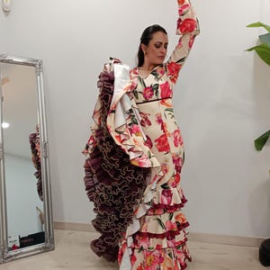 Op de afbeelding: Een crèmekleurige flamencojurk met een bloemenpatroon in rood en roze. De jurk heeft meerdere gerimpelde lagen en lange mouwen met uitlopende manchetten. Een vrouw draagt de jurk en heeft haar arm omhoog.