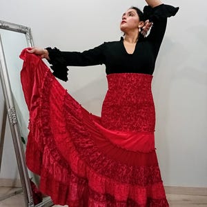 Op de afbeelding: Een flamencodanseres in een zwarte top met ruchesmouwen en een felrode rok. De rok heeft meerdere lagen stof en ruches, wat een vloeiend effect creëert. De armen van de danseres zijn geheven, wat het ontwerp van het kostuum benadrukt.