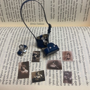 Könnte beinhalten: Ein blaues Lederkameraband mit einer Miniaturkamera und mehreren alten Fotos, die daran befestigt sind. Die Fotos zeigen ein junges Mädchen in einer Balletpose, ein junges Mädchen in einem Kleid und einen jungen Jungen im Anzug.