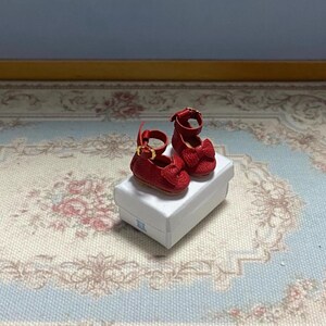 May include: Miniature red Mary Jane shoes with bow accents and gold-colored buckles, displayed on a white box. The shoes are a vibrant red, showcasing a classic design. The box sits on a floral patterned surface.