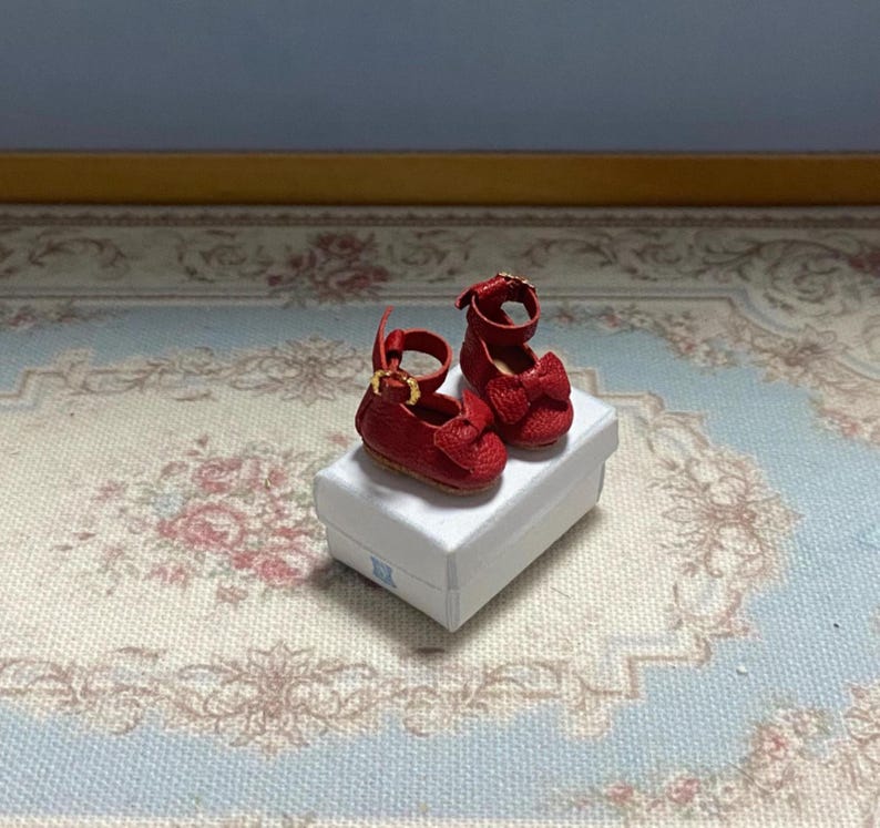 May include: Miniature red Mary Jane shoes with bow accents and gold-colored buckles, displayed on a white box. The shoes are a vibrant red, showcasing a classic design. The box sits on a floral patterned surface.
