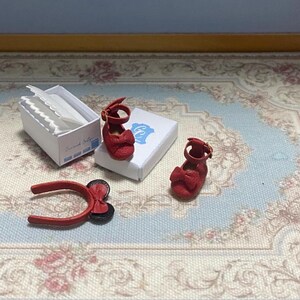 May include: Miniature red Mary Jane shoes with bow detail, a matching headband, and a small white box with a blue logo. The shoes have a strap with a buckle. The items are arranged on a floral patterned surface.