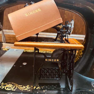 Può includere: Una macchina da cucire Singer vintage con custodia marrone. La macchina è nera con scritte dorate e dettagli ornati. La custodia è aperta, rivelando i componenti della macchina. La parola "SINGER" è visibile sulla custodia e sulla base della macchina.