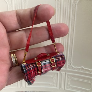 May include: A small red and black plaid bag with a red leather strap and gold buckles. The bag is about the size of a hand.