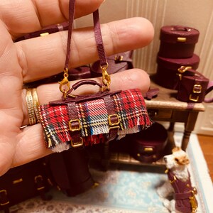 Puede incluir: Un bolso de mano de cuero burdeos en miniatura con una solapa de tela a cuadros y hebillas doradas. El bolso tiene una larga correa de cuero burdeos con cierres dorados.