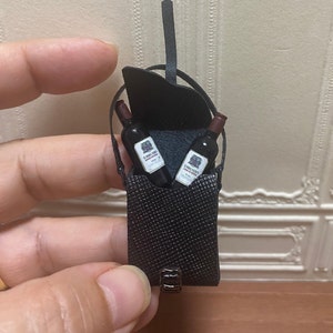 Puede incluir: Un porta vinos de cuero negro con una correa para el hombro y dos botellas de vino tinto en miniatura en su interior.