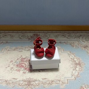 May include: A pair of miniature red doll shoes with ankle straps and bow details. The shoes are displayed on a small white box, set against a patterned rug and a light blue wall. The shoes are approximately 1 inch in length.
