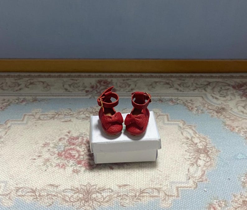 May include: A pair of miniature red doll shoes with ankle straps and bow details. The shoes are displayed on a small white box, set against a patterned rug and a light blue wall. The shoes are approximately 1 inch in length.