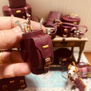 Puede incluir: Miniatura de bolsa de golf burdeos con palos de golf, herrajes dorados y equipaje a juego. El conjunto incluye una pequeña figura de perro con abrigo y correa burdeos. También se ven otros accesorios burdeos.