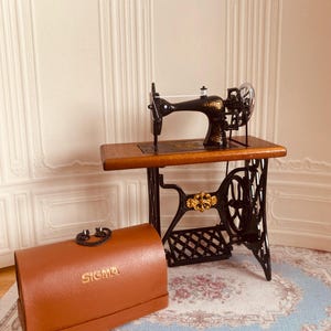 Può includere: Una macchina da cucire vintage nera con un tavolo in legno e una scatola da cucito in pelle marrone con la scritta "SIGMA" in rilievo. La macchina da cucire è su un tappeto con motivi bianchi e blu.