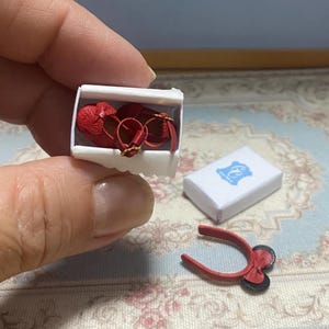 May include: Miniature red heart-shaped shoes with gold buckles inside a white box. A small white box with a blue emblem and a red bow-shaped accessory are also present. The items are likely for a doll or a craft project.