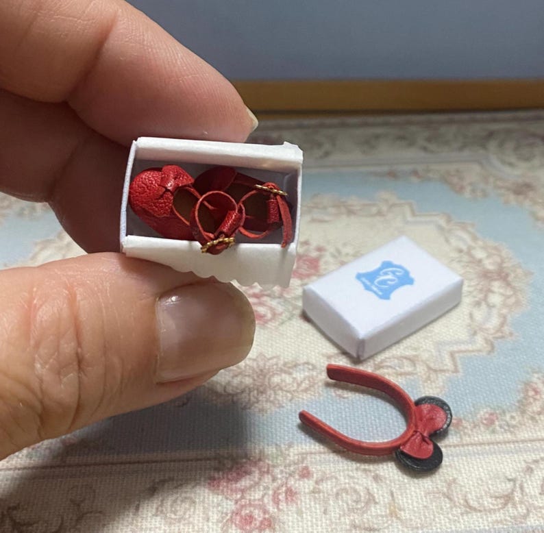 May include: Miniature red heart-shaped shoes with gold buckles inside a white box. A small white box with a blue emblem and a red bow-shaped accessory are also present. The items are likely for a doll or a craft project.